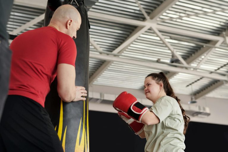 une jeune fille trisomique fait de la boxe comme sport adapté. Elle est équipée de gants de boxe et tape dans un sac de sable tenu par un homme.
